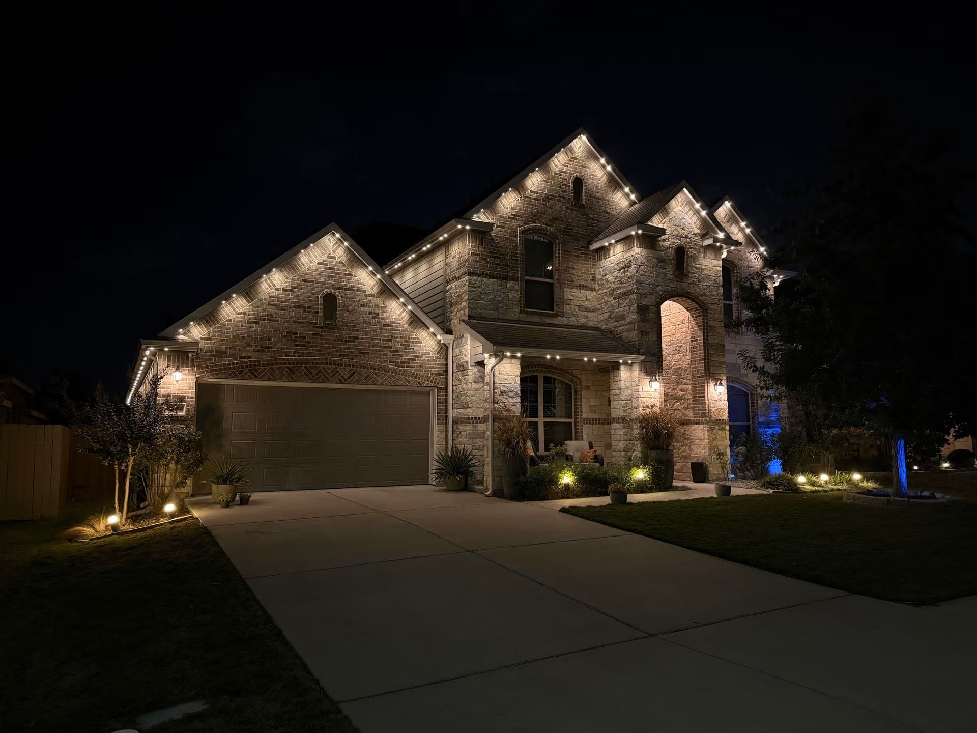 Stone house exterior at night with lit facade, garage, driveway, and landscape lighting