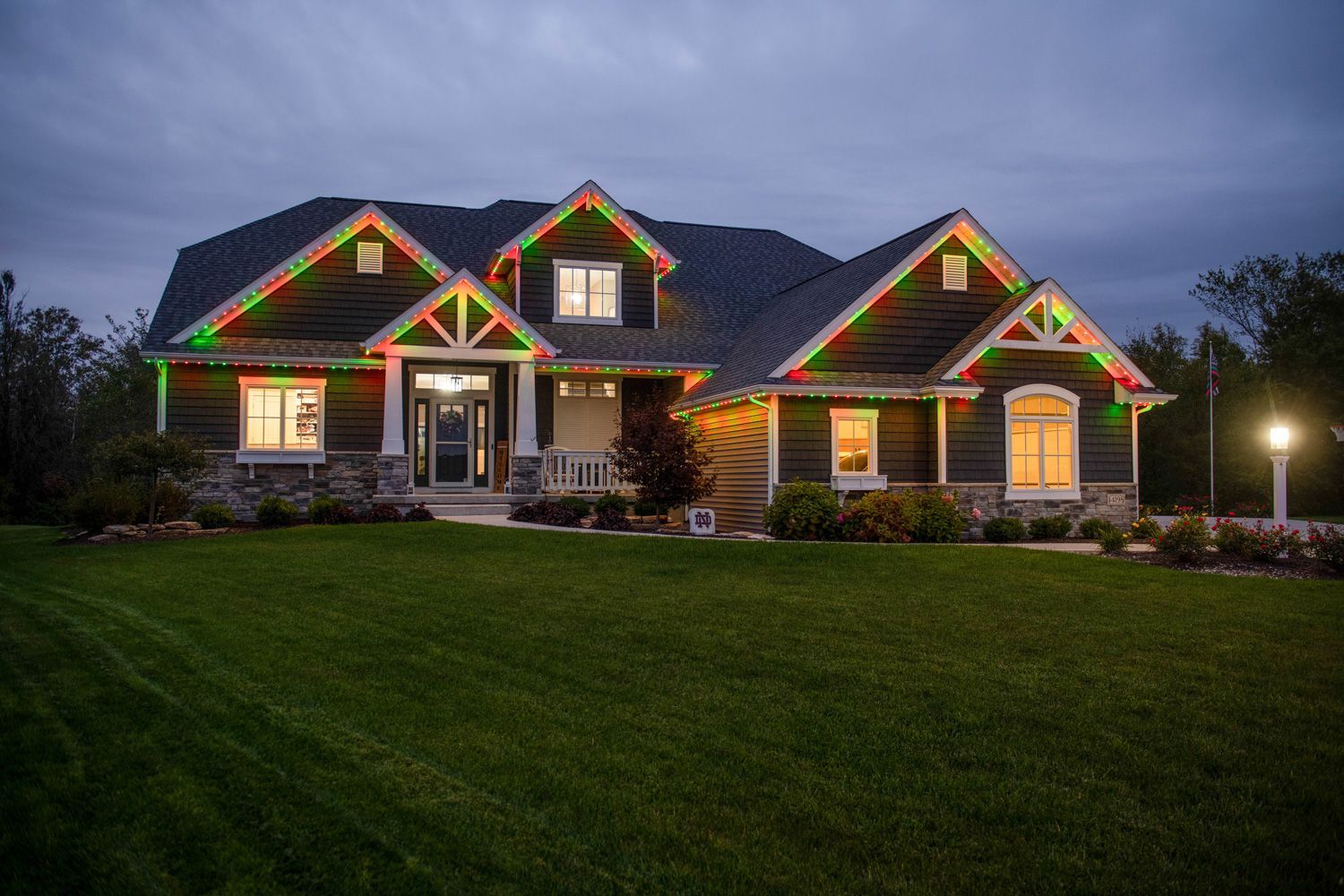 House at dusk decorated with colorful Christmas lights, glowing windows, and a green lawn