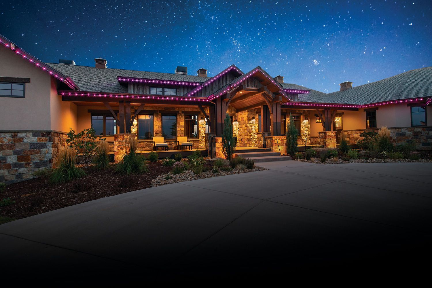 Stone house at dusk with warm glowing lights, pink roof accents, and a curved driveway under a starry sky