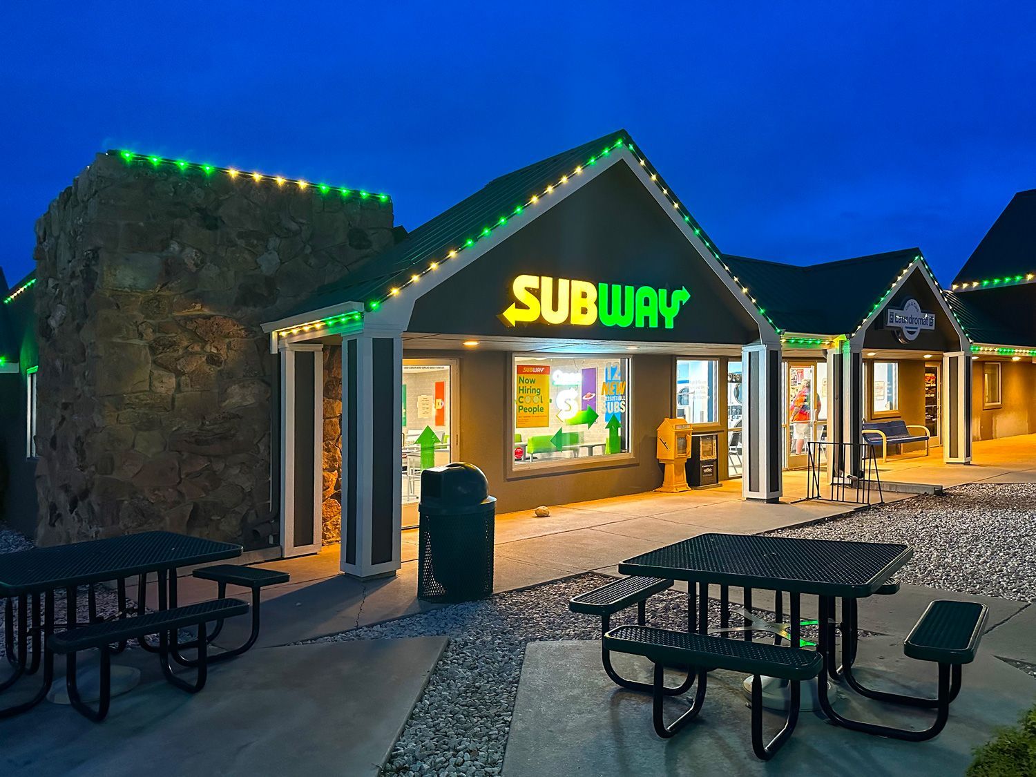 Subway restaurant storefront at dusk with green-lit roofline and outdoor picnic tables