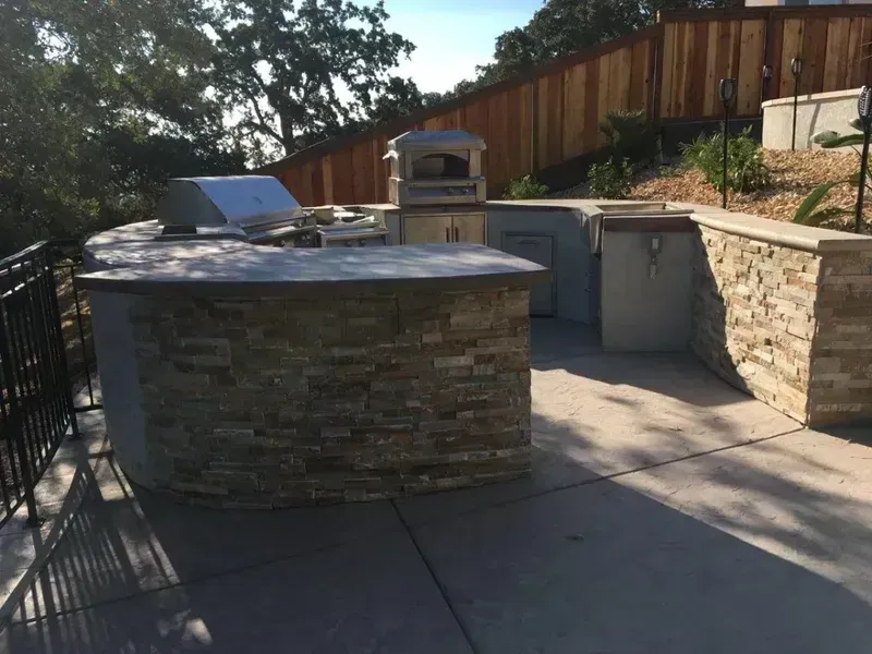 Outdoor stone patio kitchen with grill and counters in a fenced backyard