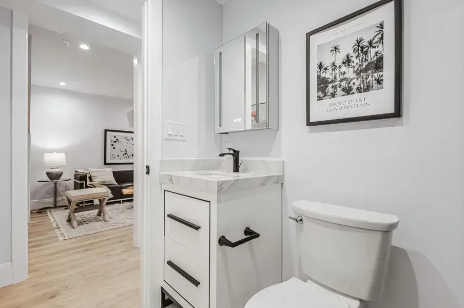 Bright white bathroom with vanity and toilet, opening to a light living room with wood floors