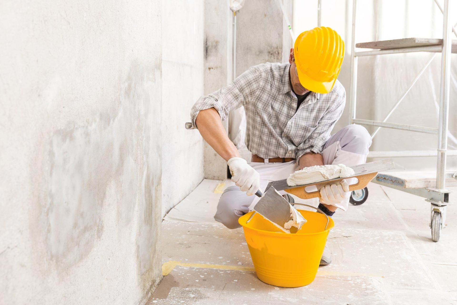 A man is kneeling down next to a yellow bucket and a spatula.