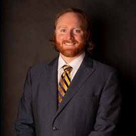 A man with red hair and a beard, wearing a navy suit and a yellow and navy striped tie, smiling against a dark background.