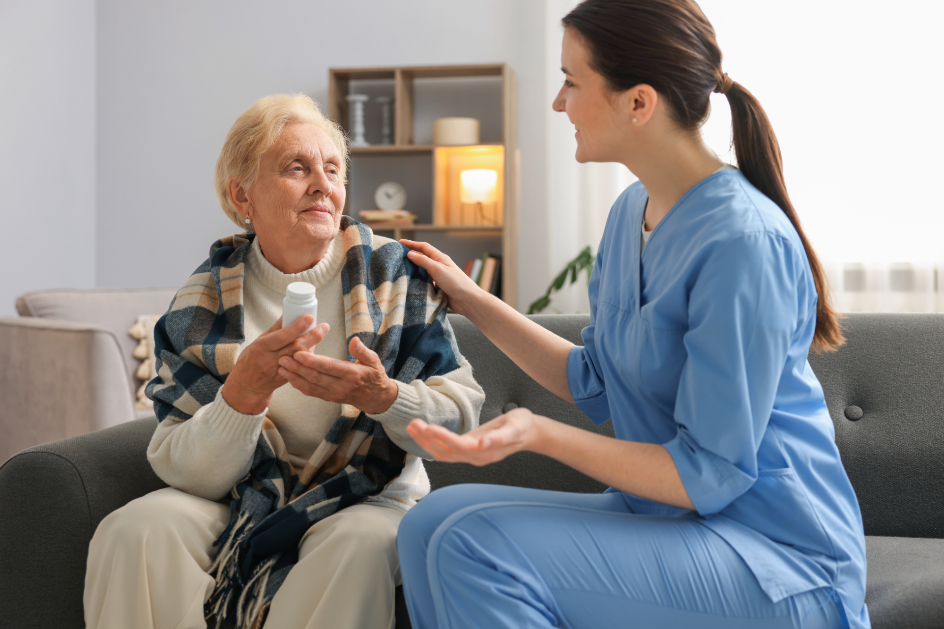 A caregiver in blue scrubs talks with an elderly person holding a bottle of pills while sitting on a couch.