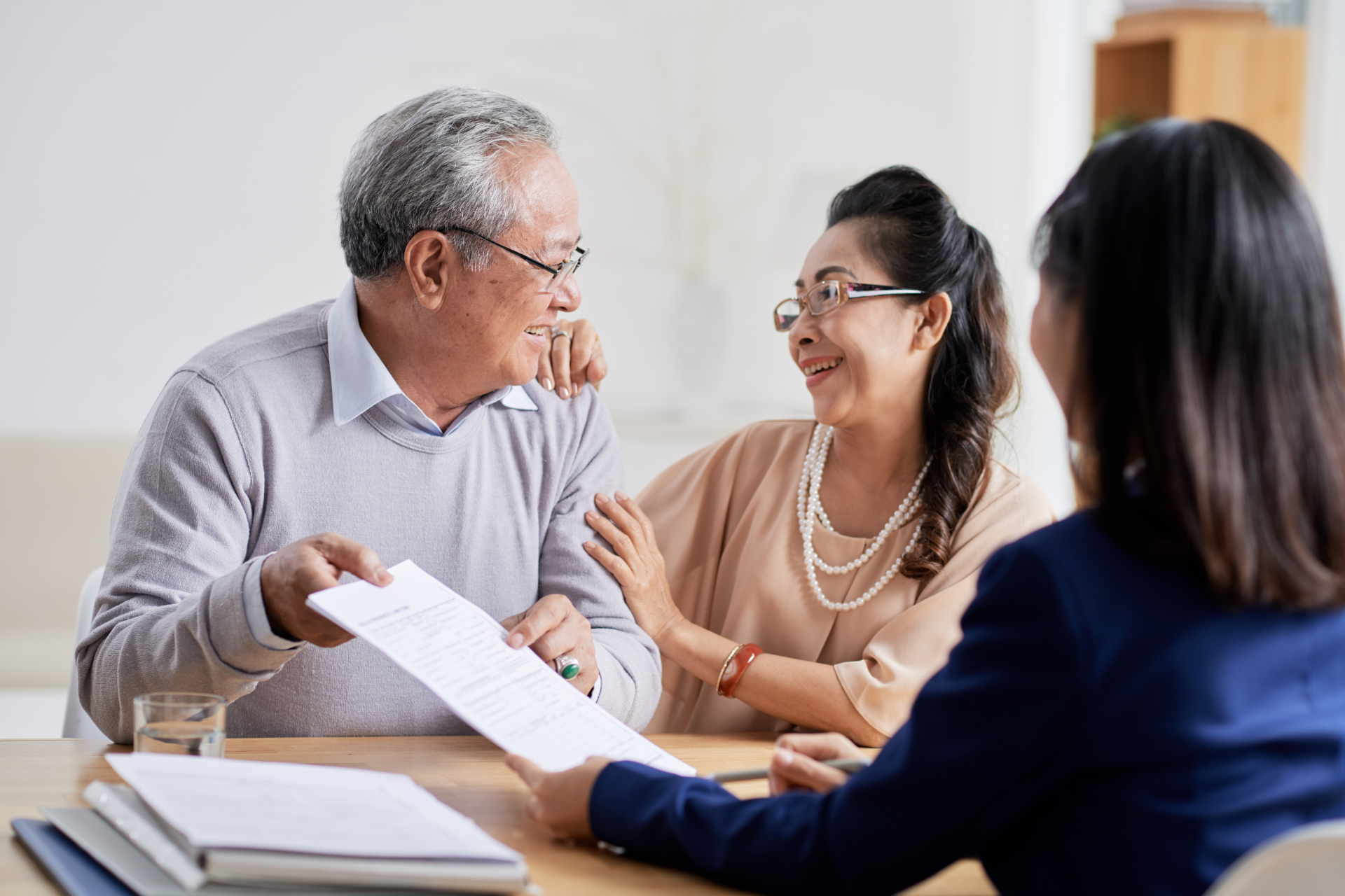 An elderly couple discusses documents with a professional sitting across a wooden table in a bright office.