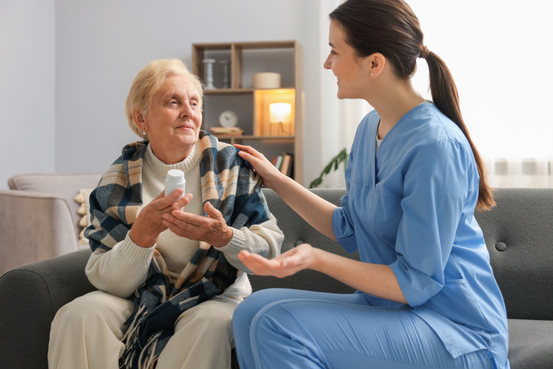A caregiver in blue scrubs talks with an older person sitting on a sofa, who is holding a medicine bottle.