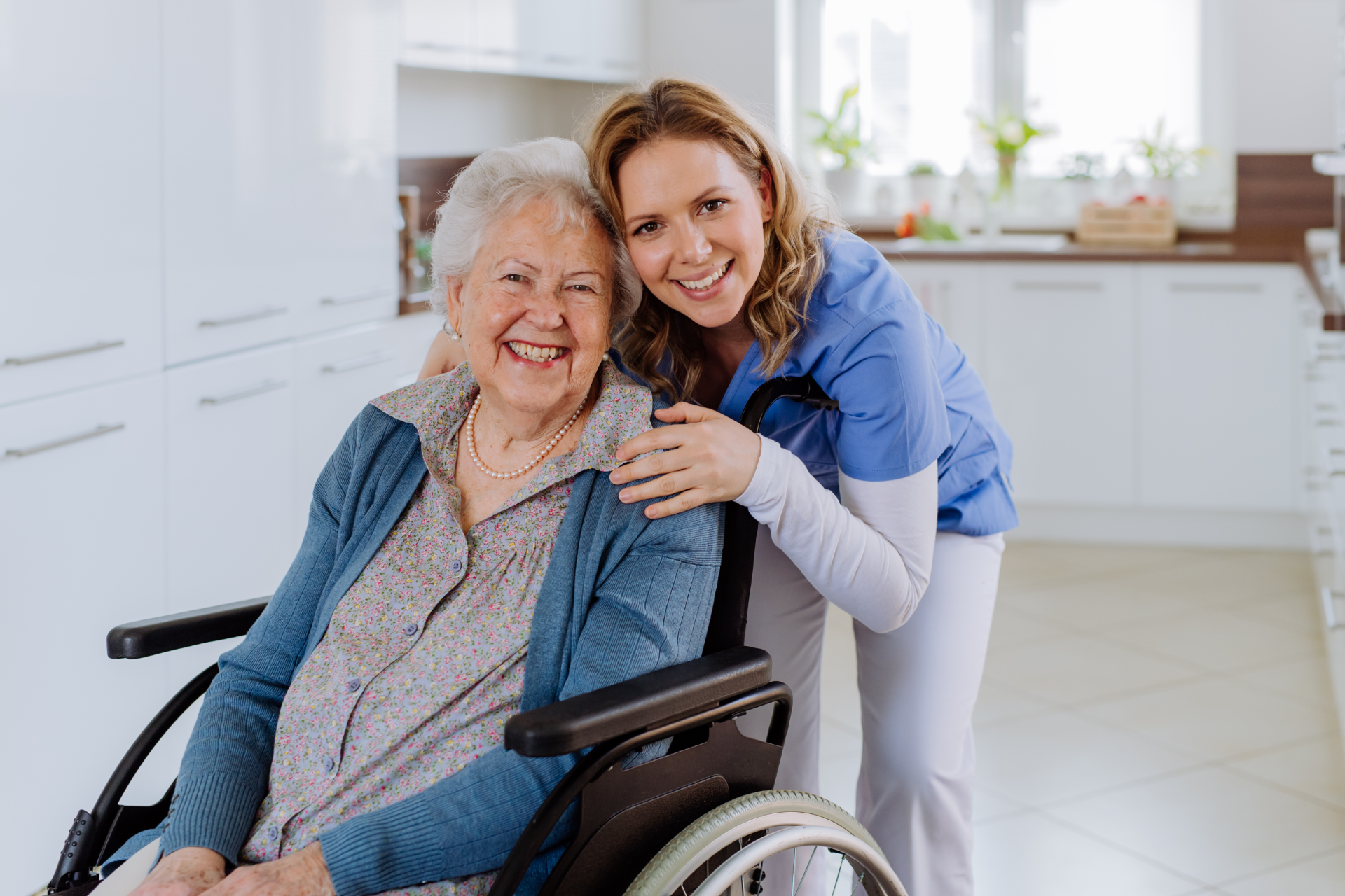 A caregiver in blue scrubs smiles while resting a hand on the shoulder of a seated person in a wheelchair in a kitchen.