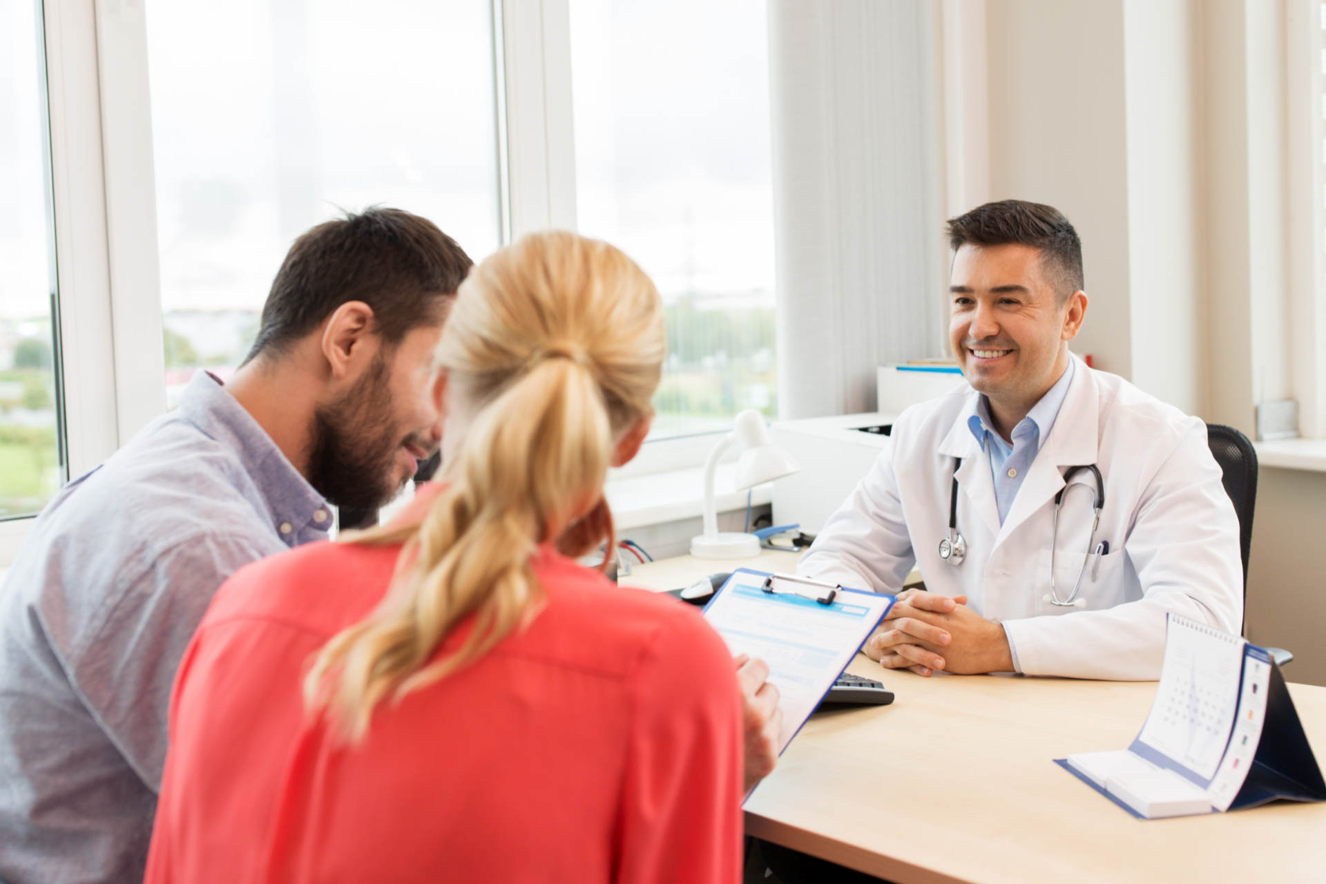 A doctor in a white coat sits at a desk talking to a couple in a brightly lit office.