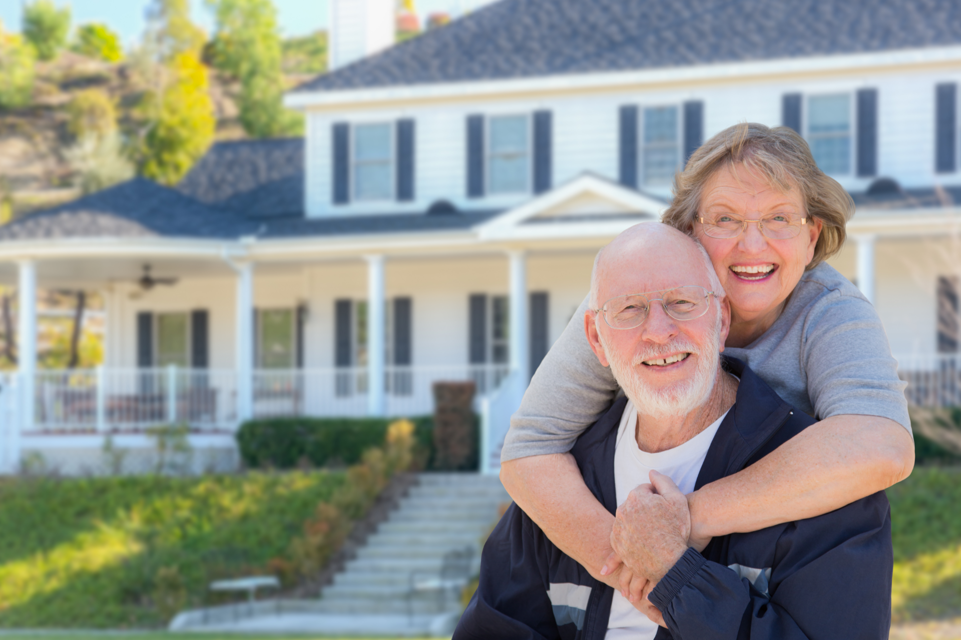 A smiling couple embraces outdoors in front of a white two-story house with a porch and green lawn.