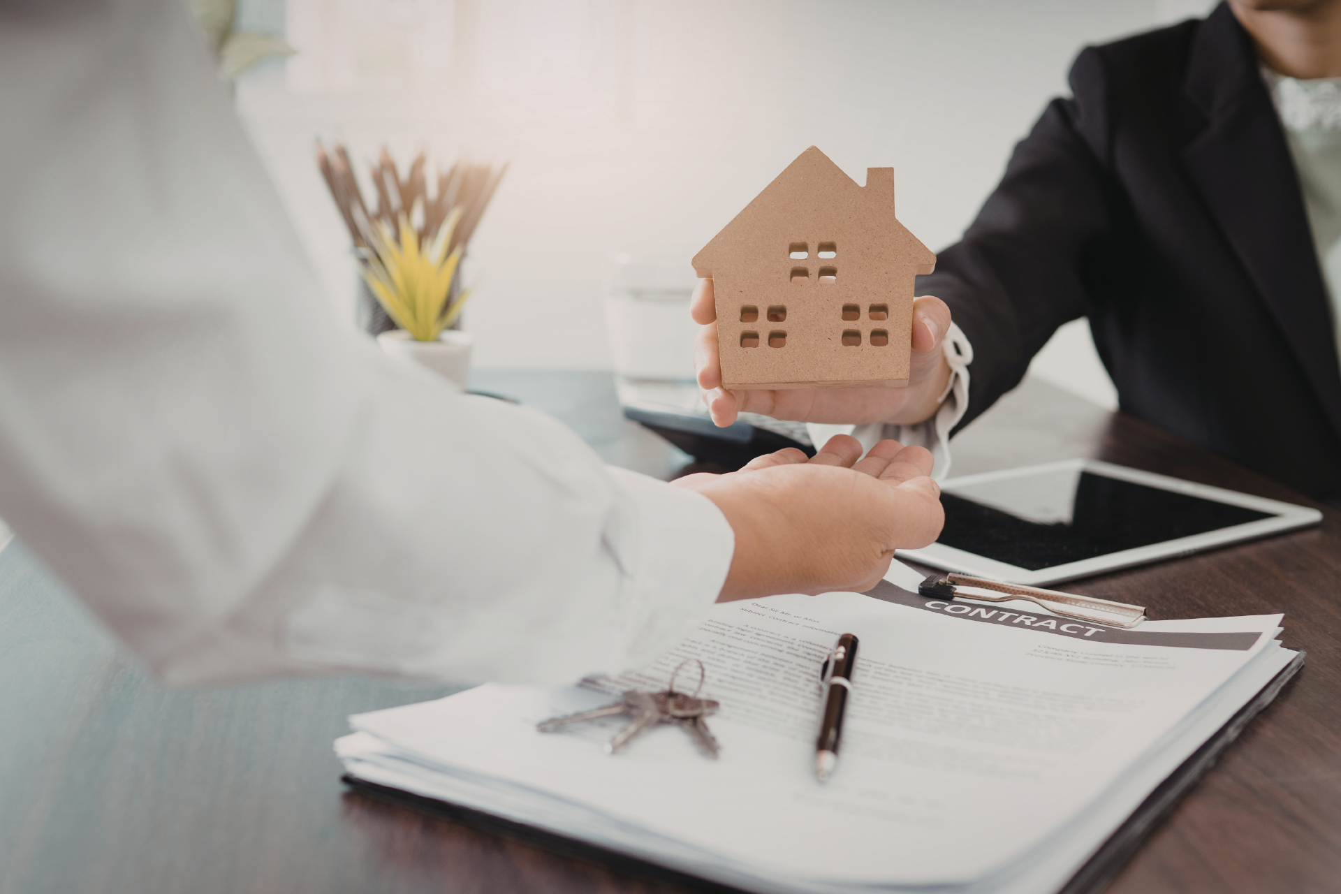 A professional in a suit hands a wooden house model to a client across a desk with a contract and keys.