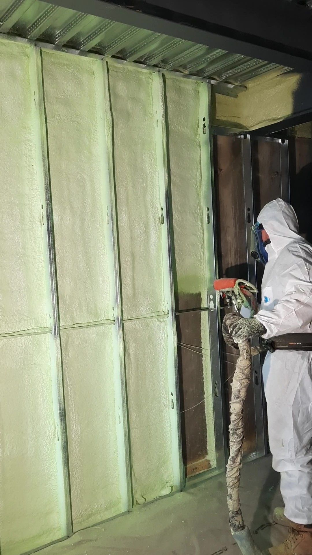 Une personne en combinaison de protection pulvérise de la mousse isolante sur un mur à l'intérieur d'un bâtiment.