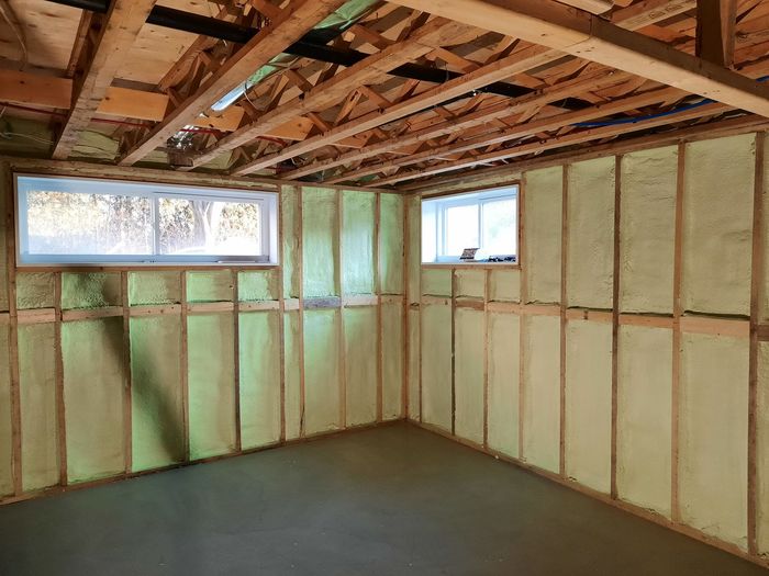 Vue intérieure d'un sous-sol avec isolation en mousse pulvérisée entre des montants en bois et deux fenêtres.