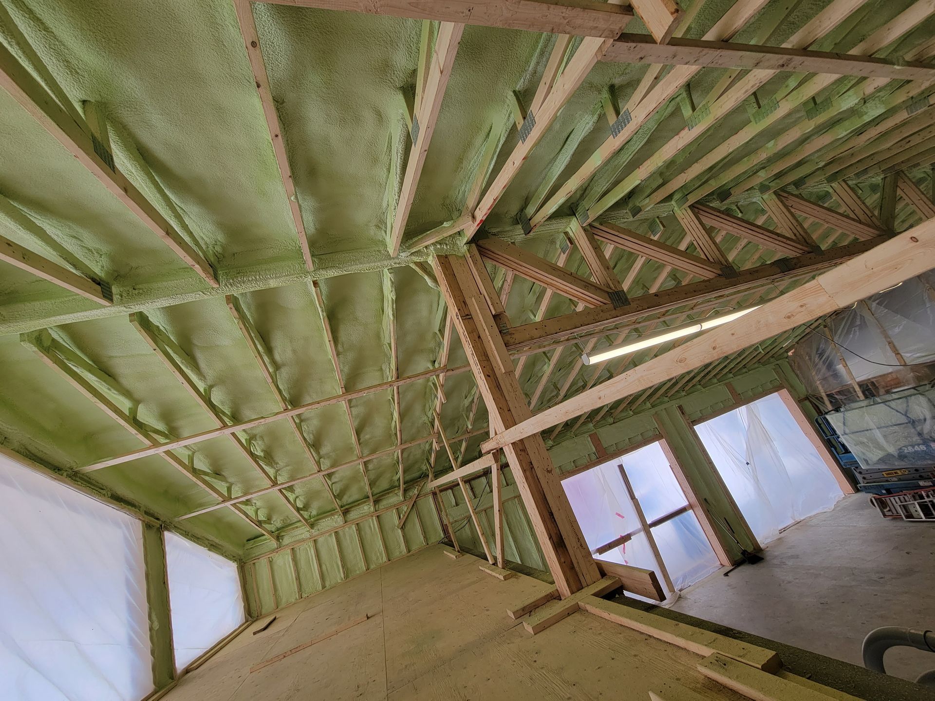 Vue intérieure d'un bâtiment en construction avec isolation en mousse projetée au plafond. Poutres et chevrons en bois visibles.