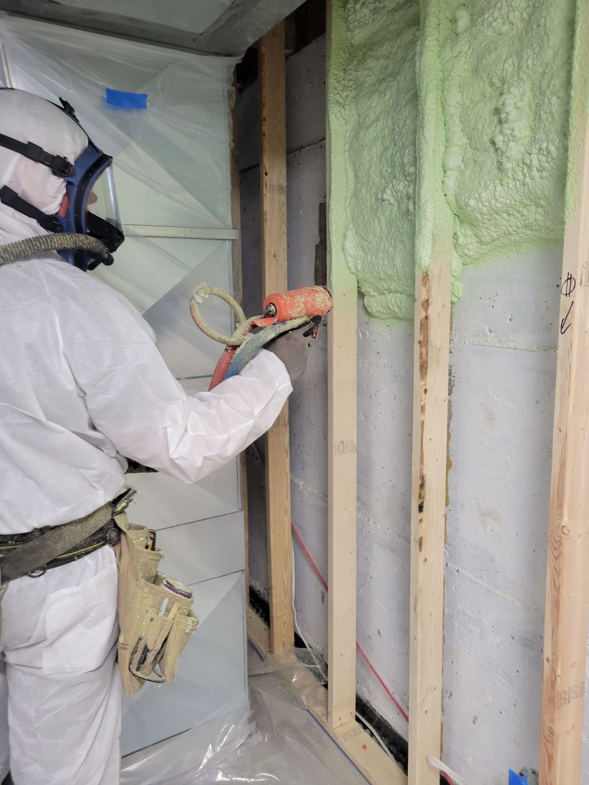 Une personne en équipement de protection pulvérise de la mousse isolante sur un mur avec des montants en bois, dans un espace confiné.