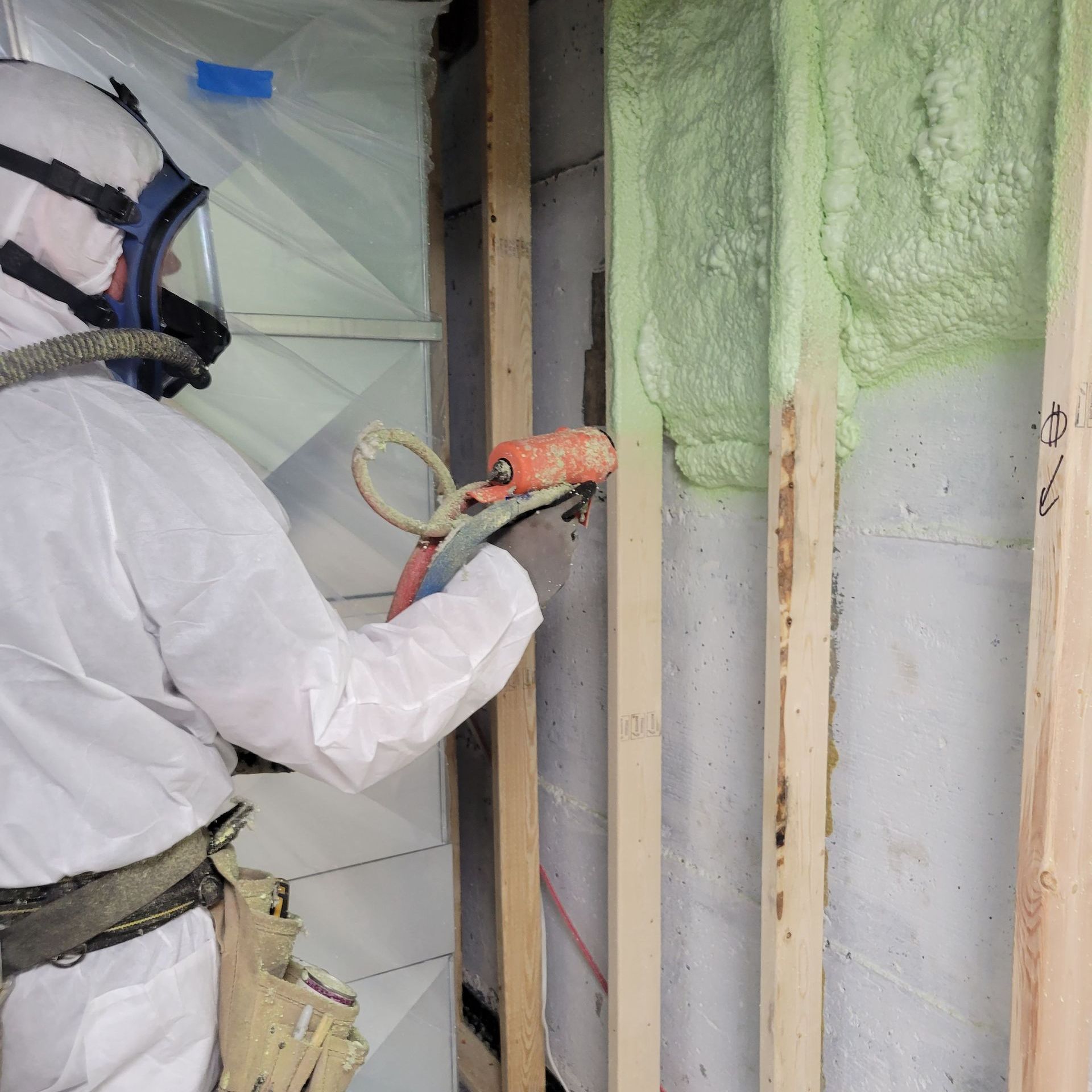 Une personne en combinaison de protection pulvérise de l'isolant en mousse verte entre des montants en bois sur un mur.