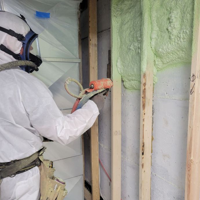 Une personne en combinaison de protection pulvérise de l'isolant en mousse verte entre des montants en bois sur un mur.