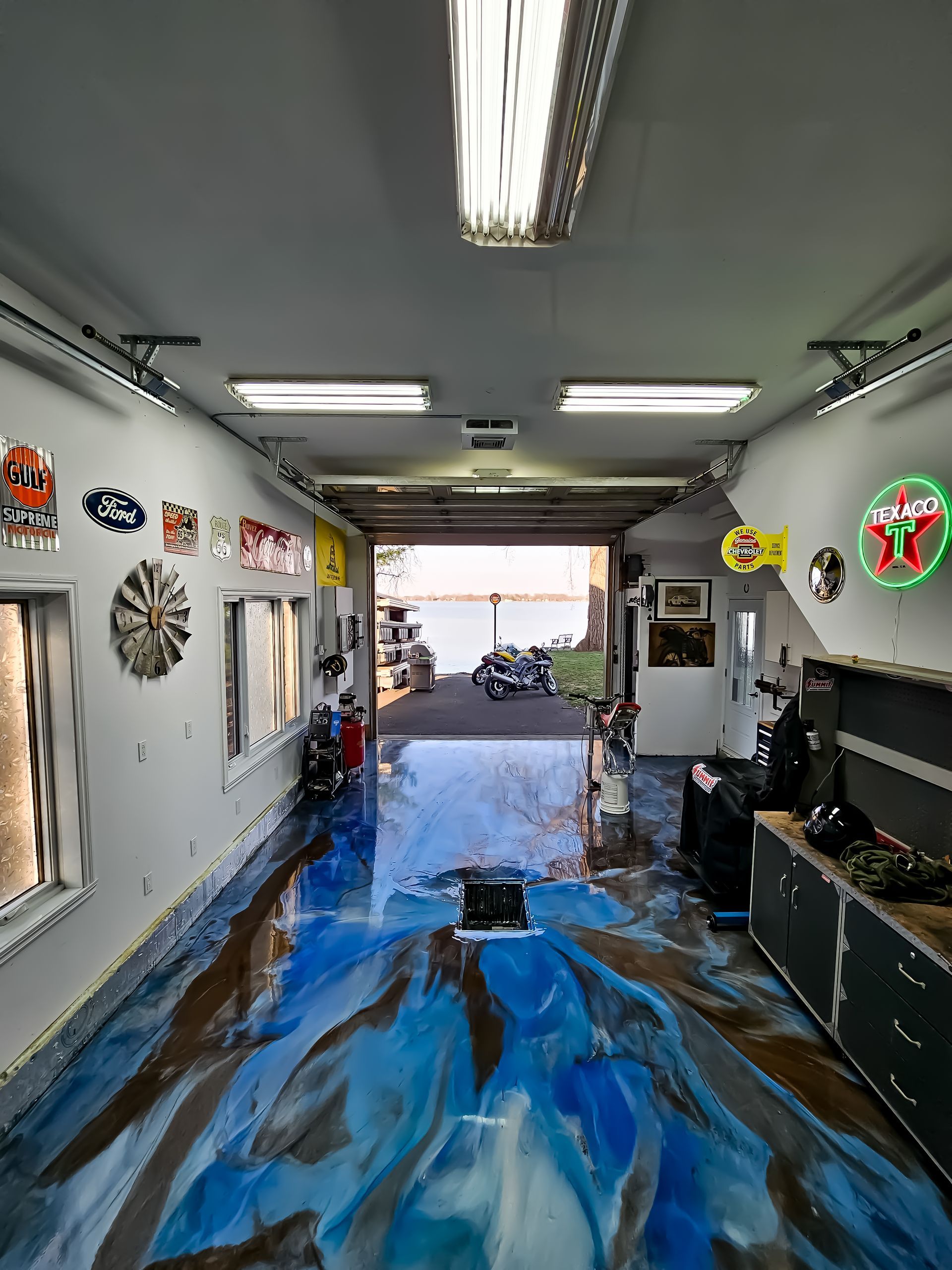 Garage avec plancher époxy bleu et marron brillant, ouvert sur une vue sur l'eau, bordé d'enseignes et de lumières.