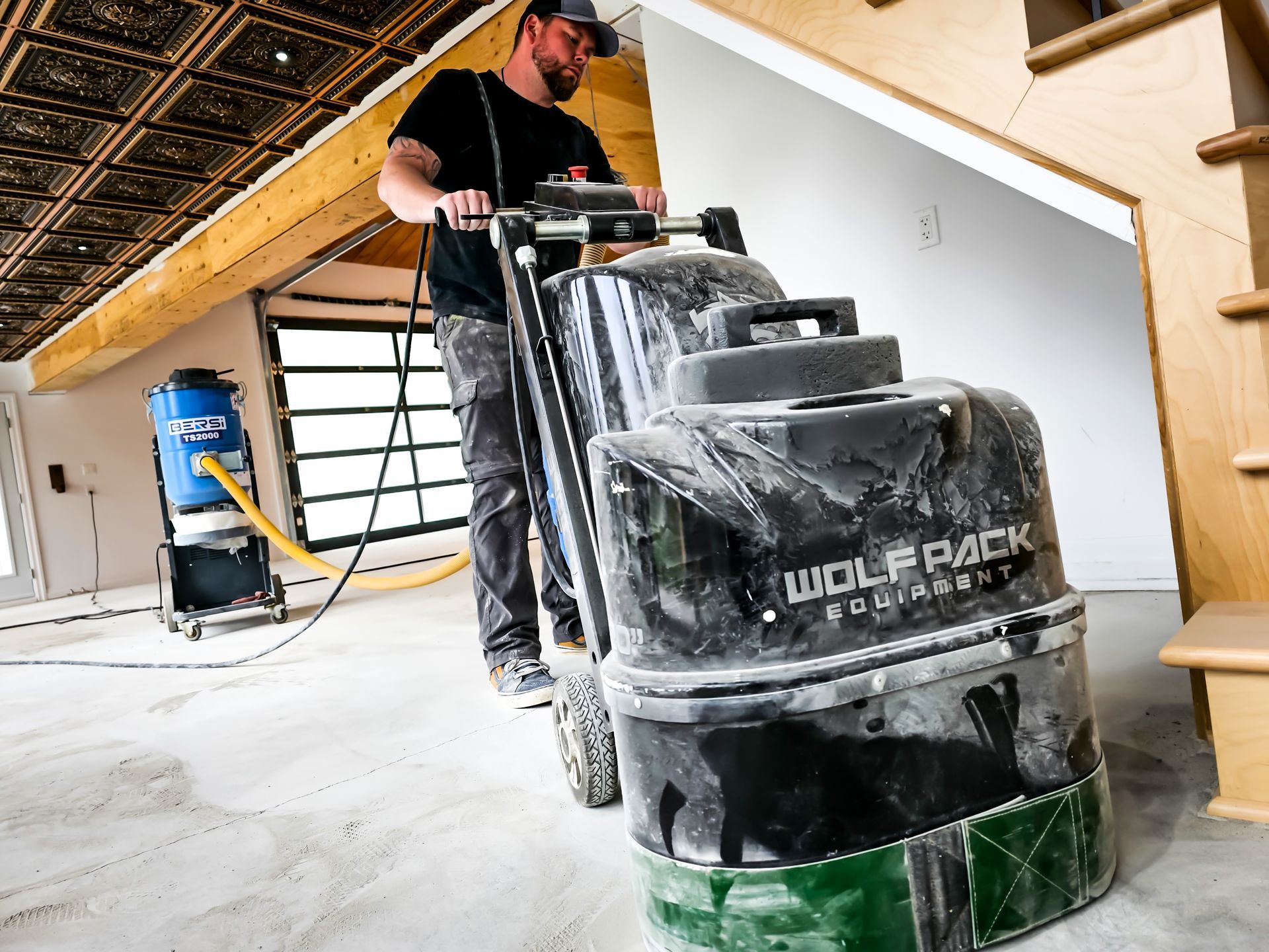 Un homme utilise une ponceuse à béton à l'intérieur ; un gros aspirateur est également présent.