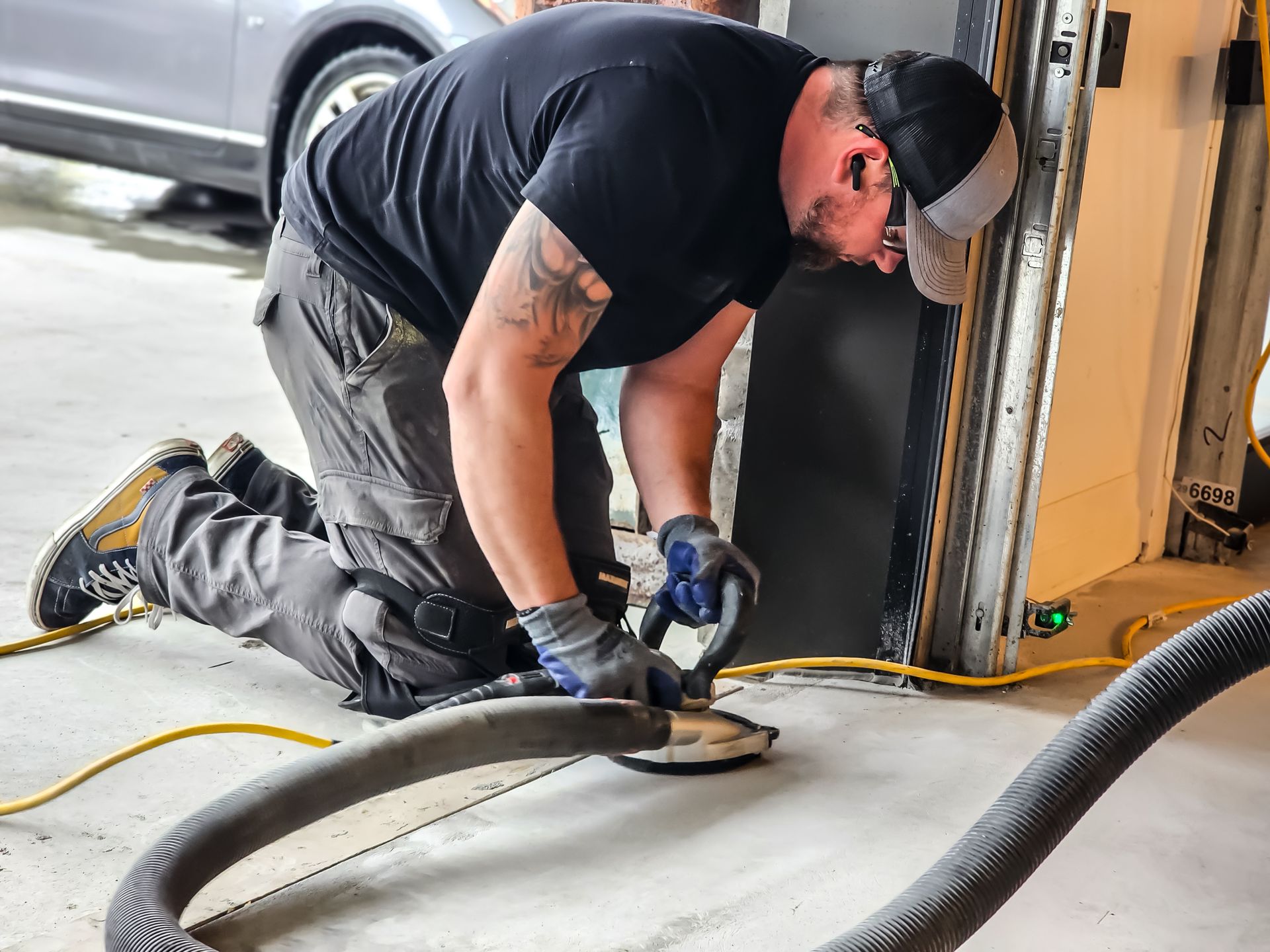 Un homme à genoux, ponce le sol en béton dans un garage, portant des gants de travail, une chemise noire et une casquette.