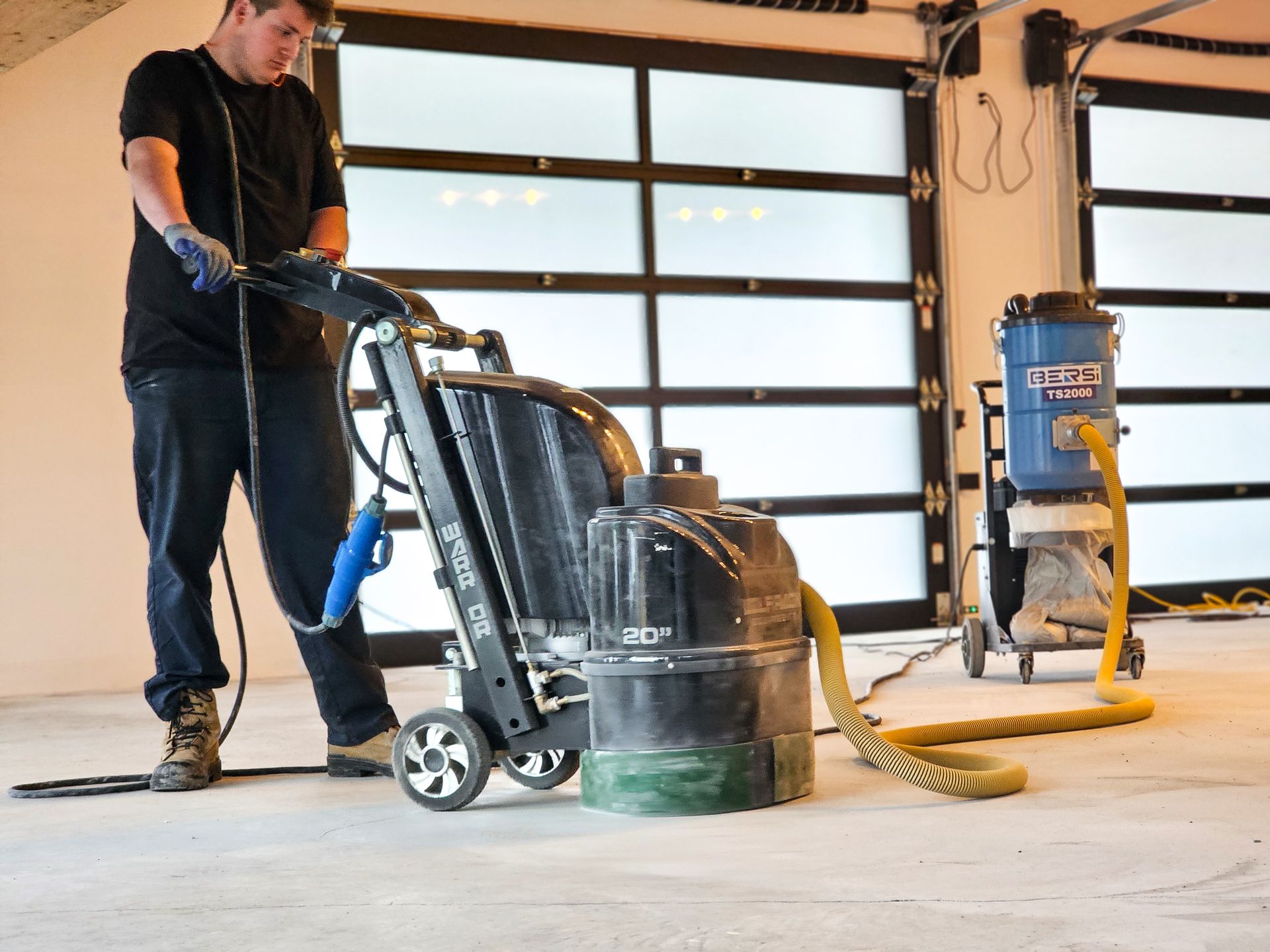 Un homme ponce un sol en béton avec une machine, reliée à un aspirateur, dans un garage.