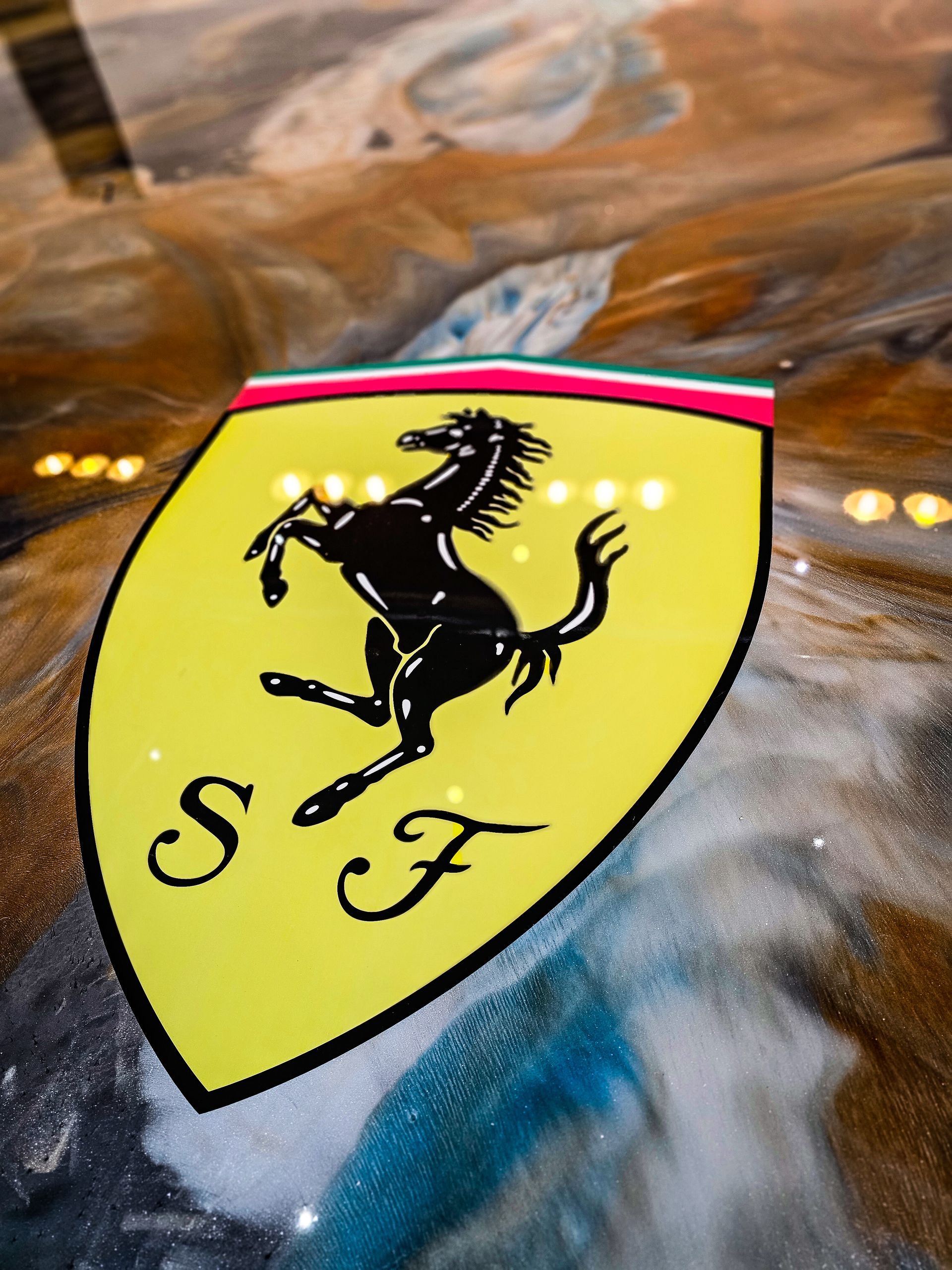 Emblème Ferrari jaune sur fond tourbillonnant bleu, marron et blanc. Le cheval cabré et la mention « SF » sont visibles.