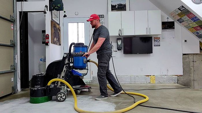 Un homme ponce un sol en béton dans un garage, à l'aide d'une machine avec un réservoir bleu, portant une casquette rouge.