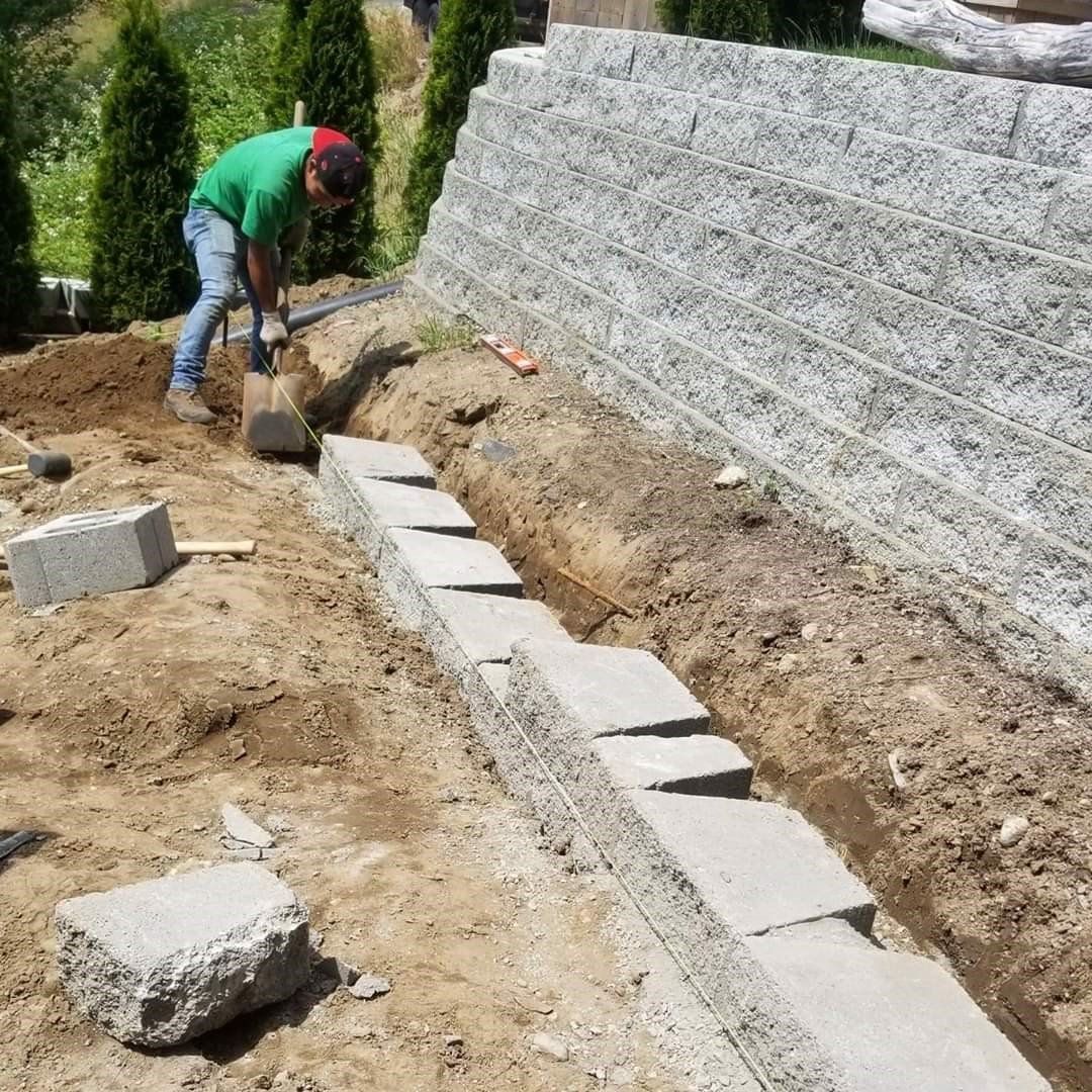 Man digging trench near a retaining wall. Blocks are laid in the trench; green shirt, outdoors.