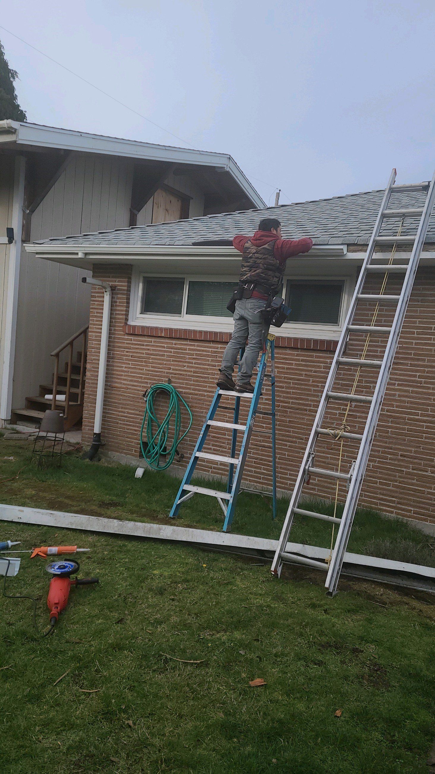 Person on a ladder working on a gutter. Red brick house, overcast sky, tools on the lawn.