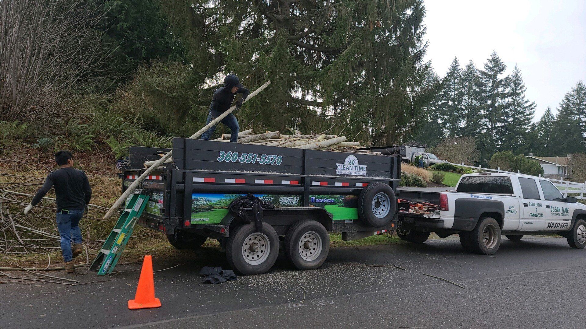 Two workers loading tree branches into a trailer, a pickup truck is parked next to them.