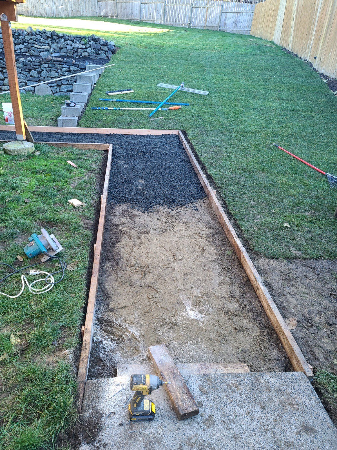 Pathway construction in backyard: wooden borders, gravel, dirt, grass. Tools and partial patio visible.