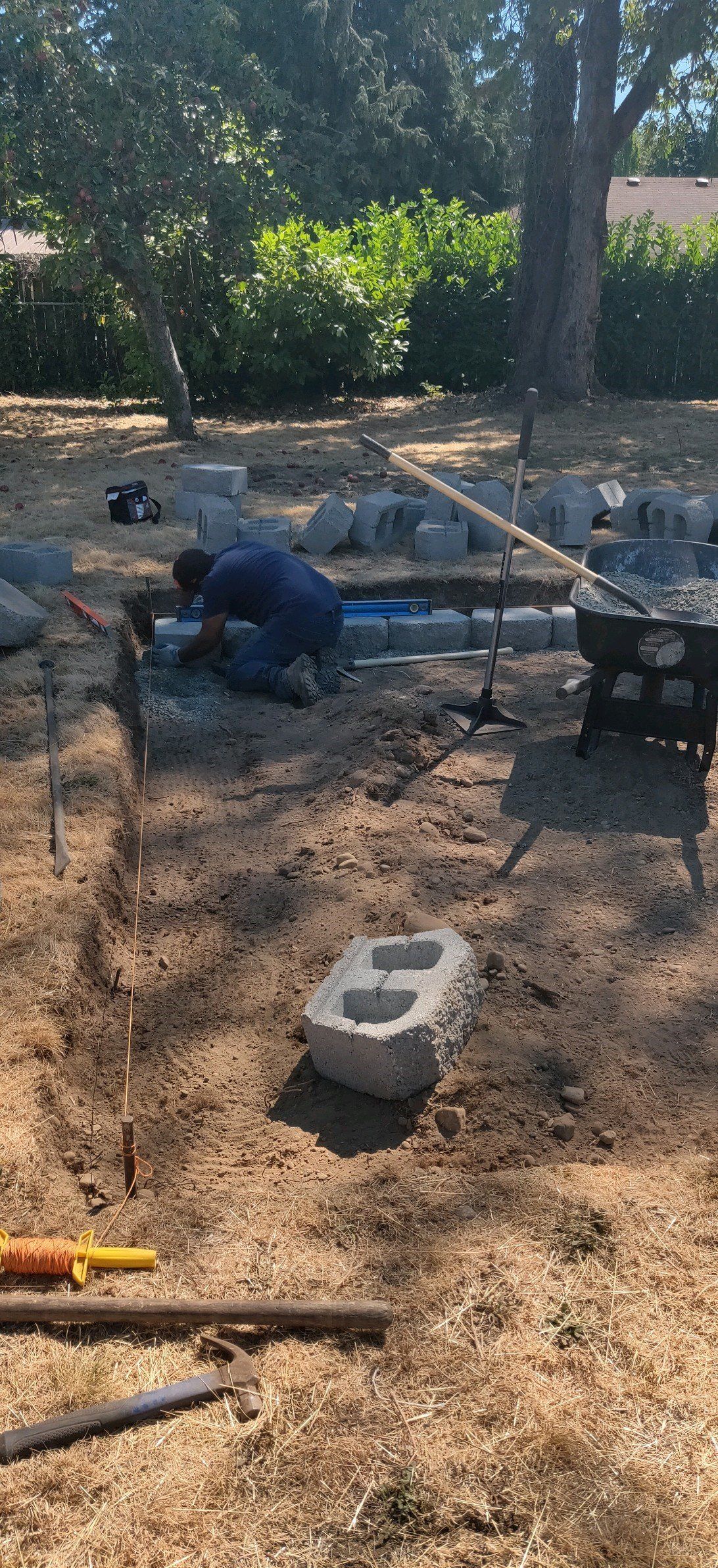 Person working on a construction project outside near trees. Cinder blocks and tools are present.