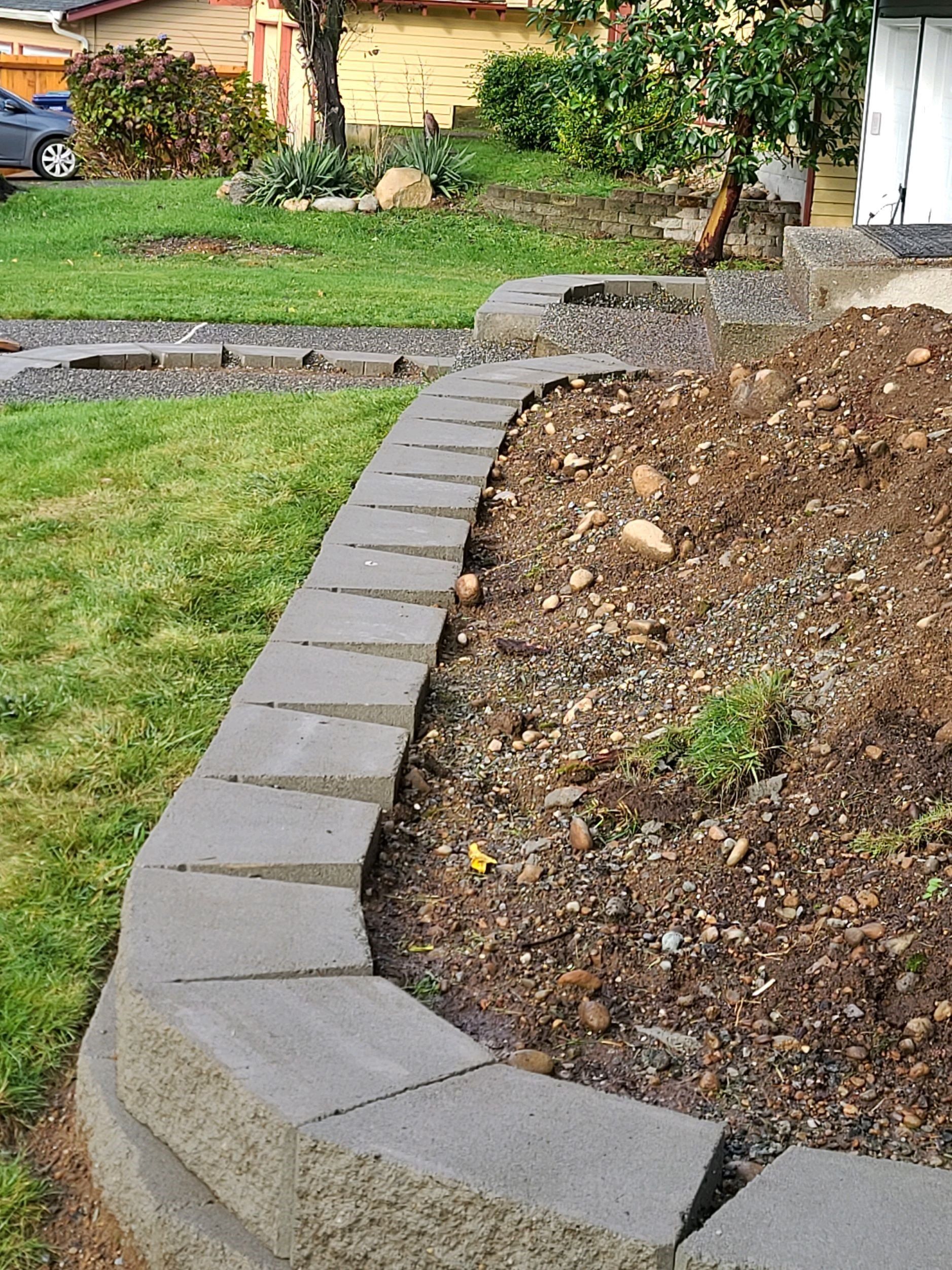 Gray retaining wall borders a brown soil flower bed in a yard with green grass.
