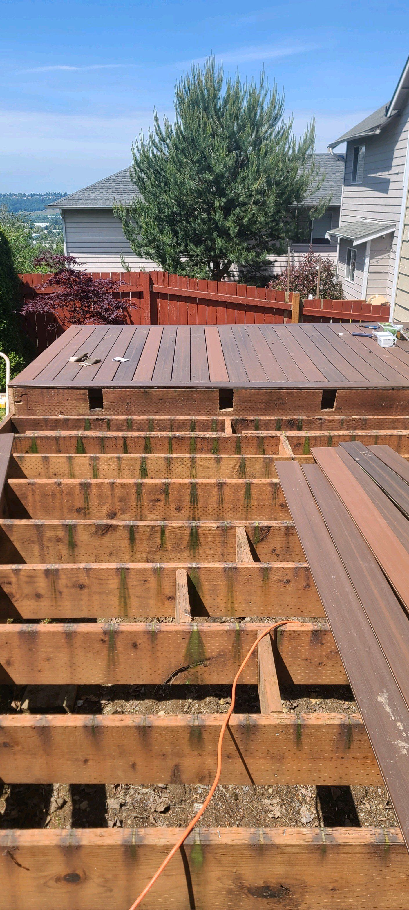 Wooden deck under construction, with exposed beams and some planks laid. A tree and houses are in the background.