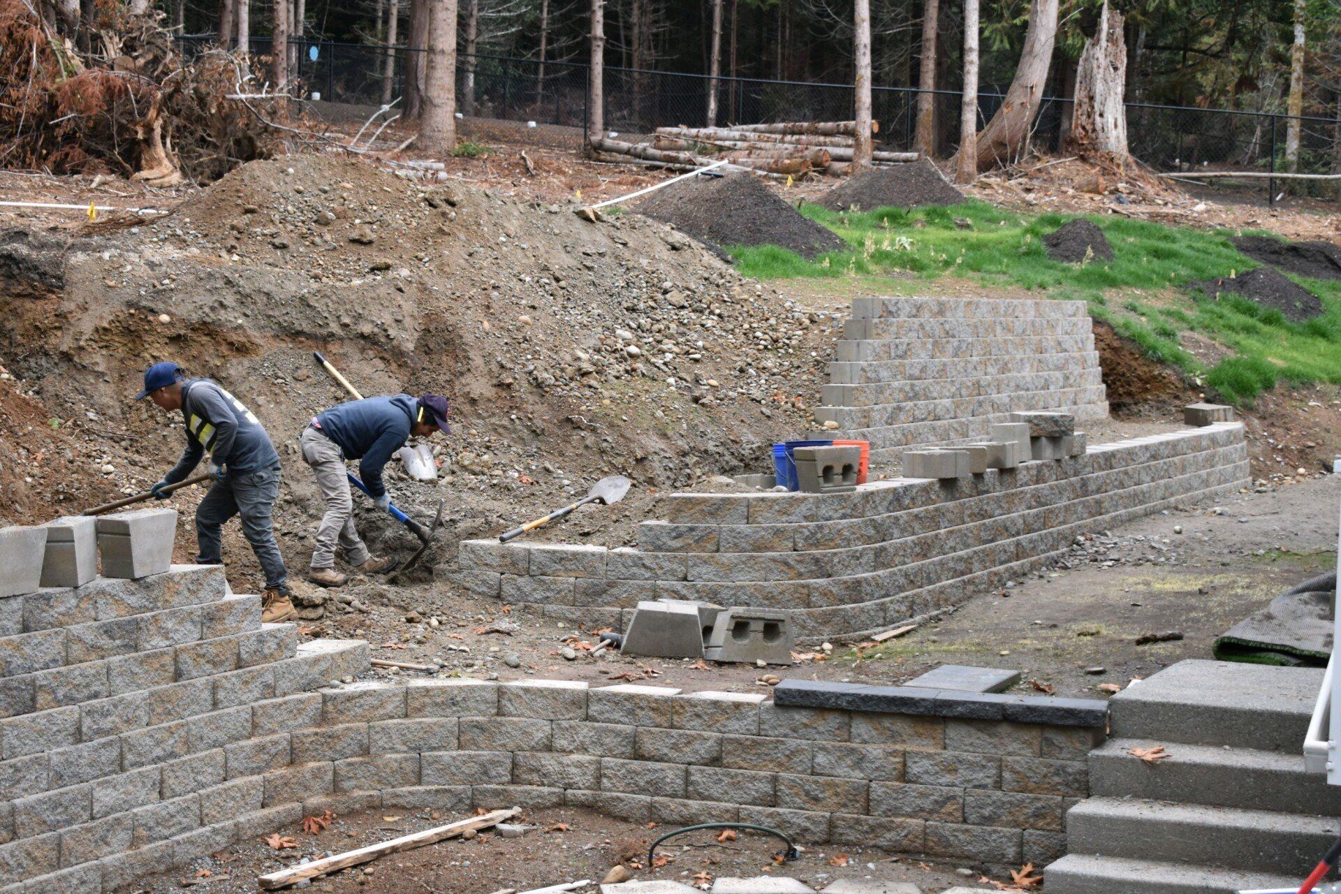 Two workers constructing retaining walls with stone blocks on a construction site.