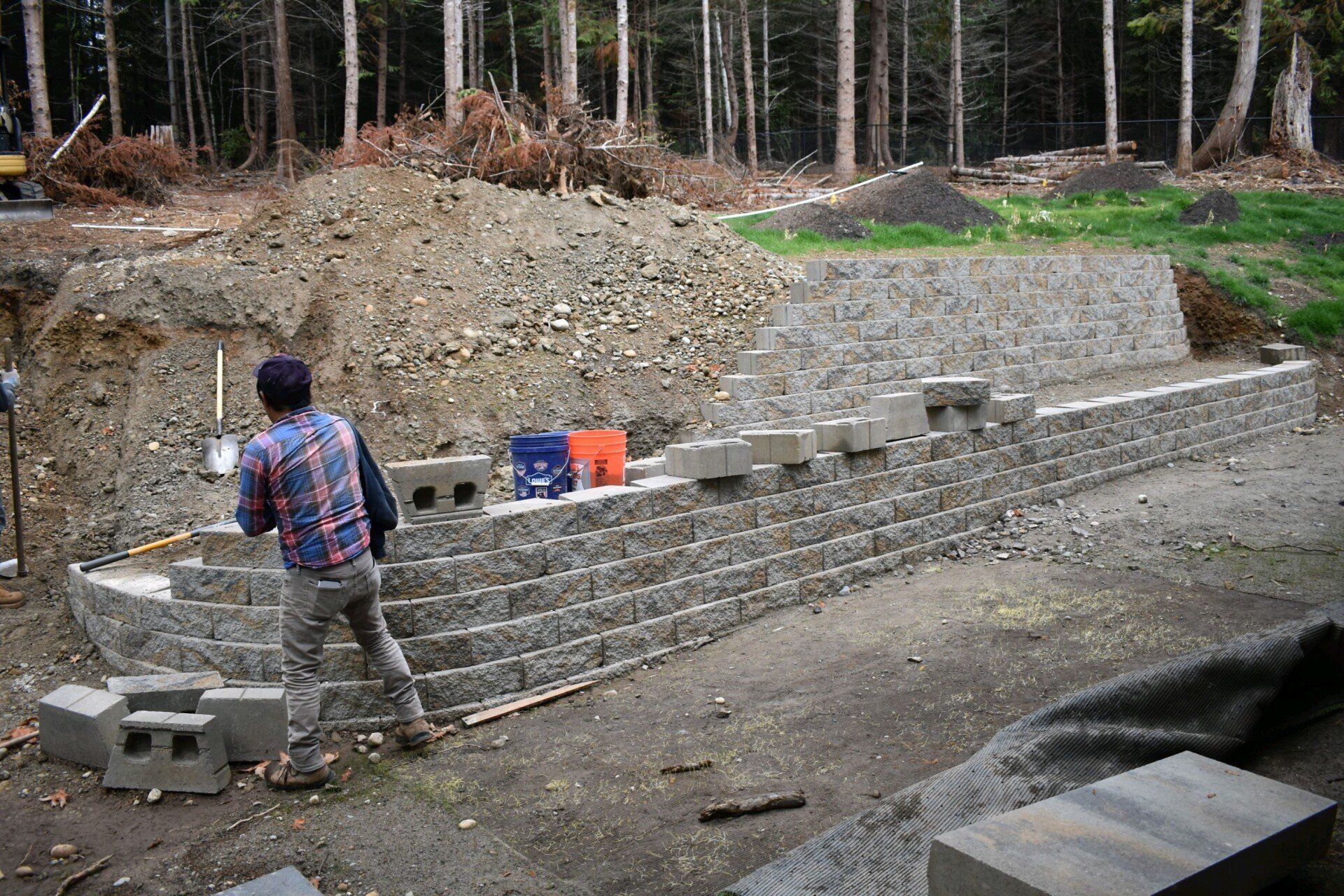 Man building a tiered retaining wall with stone blocks outdoors, in front of a forest.