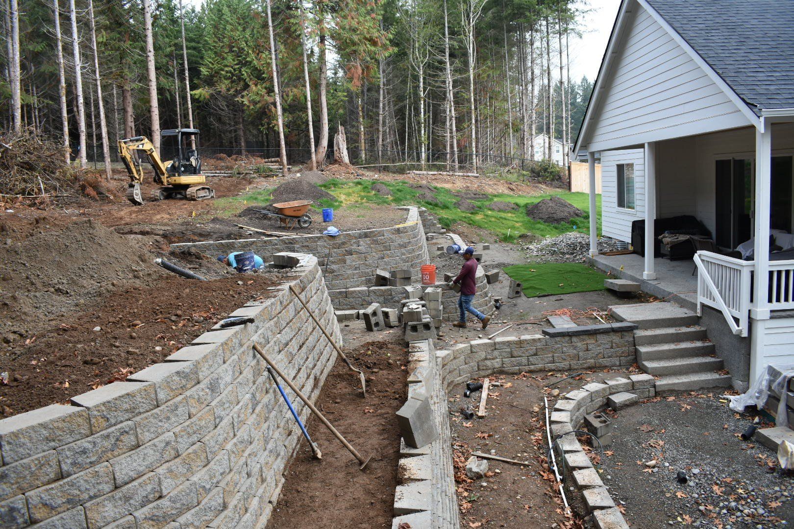 Construction site with stone retaining walls, small excavator, and workers near a house.