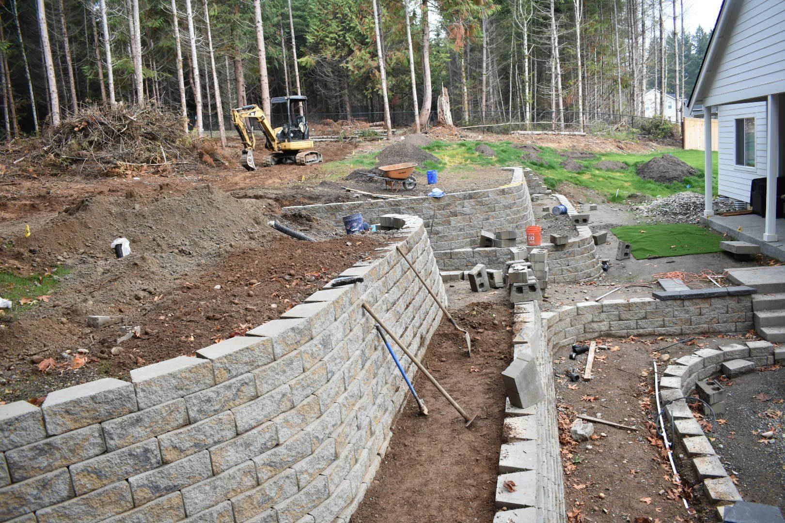 Stone retaining walls under construction in a yard, with an excavator and workers; trees in the background.