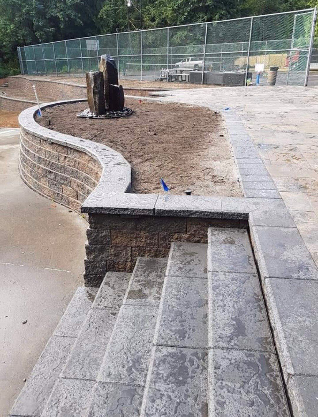 Stone steps and retaining wall leading to a raised area with a water feature, a chain-link fence in the background.
