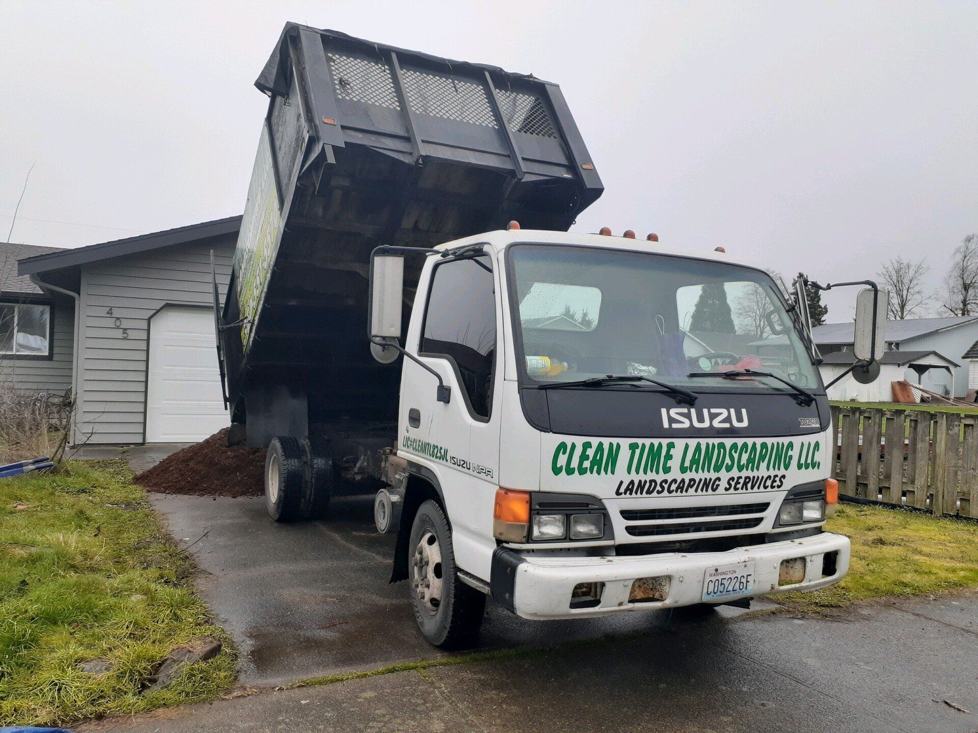 White landscaping truck dumping mulch in a driveway, next to a house. Truck reads 
