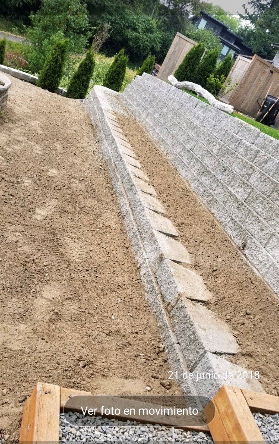 A retaining wall with stone blocks, gravel, and dirt. A few plants and trees in the background.