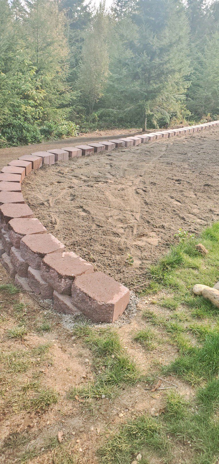 A curved retaining wall made of red bricks. Dirt inside the wall. Green grass and trees in the background.