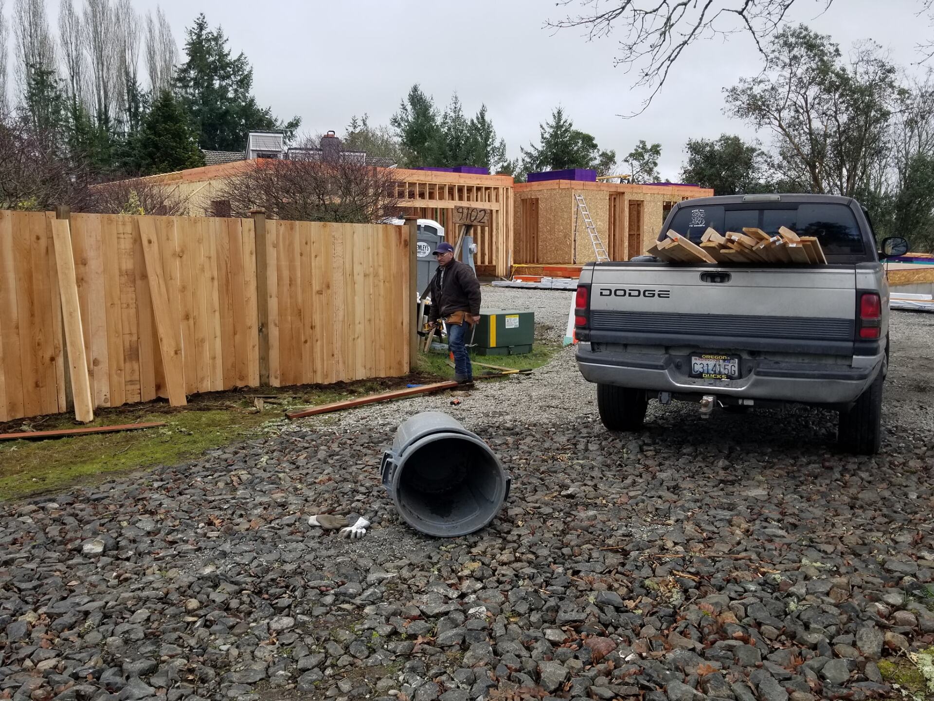 Construction site with a partially built house, wooden fence, and a parked pickup truck. A worker stands nearby.