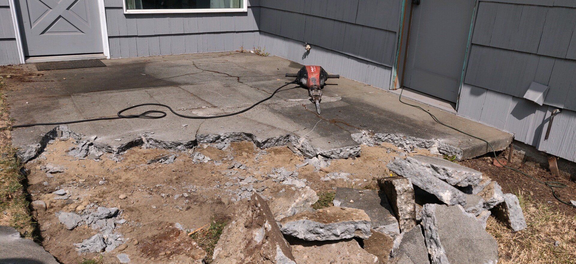 A concrete patio being demolished with a jackhammer, debris scattered nearby.