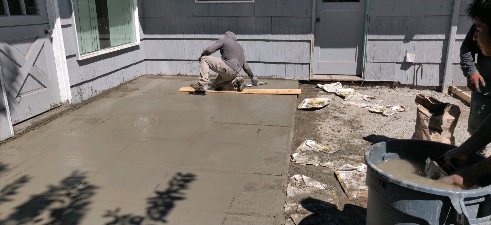 Workers pouring and smoothing concrete on a patio. One kneels, the other mixes cement in a large tub.