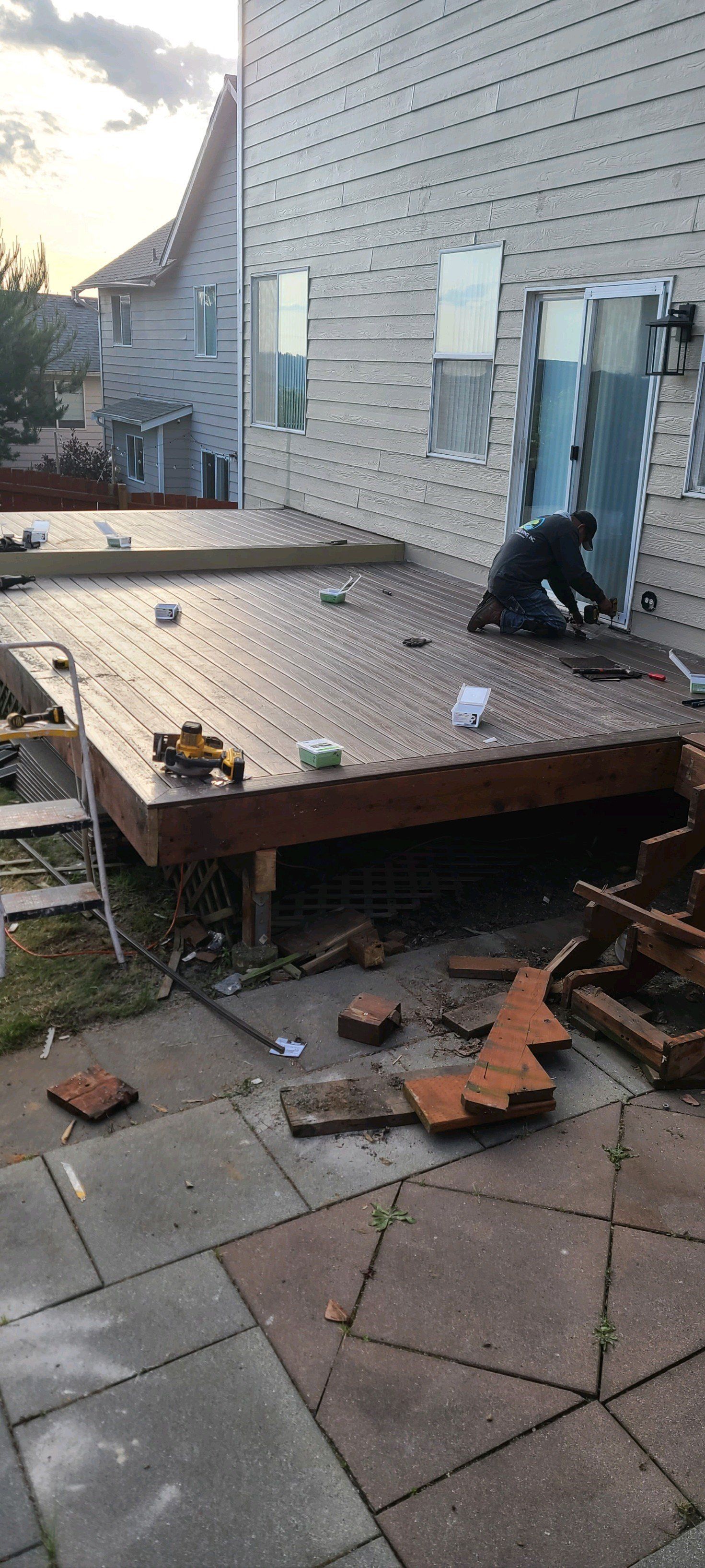 Construction worker on deck, bricks and debris on ground, gray siding, overcast sky.