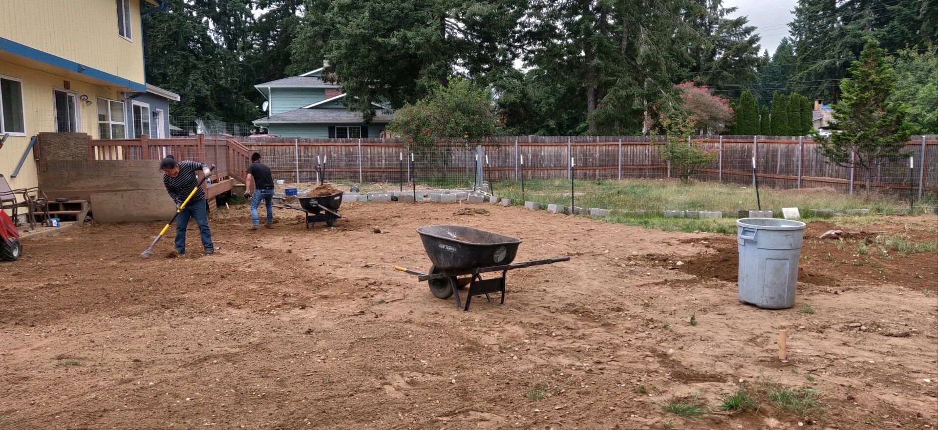 People working in a backyard, raking dirt, near a fence, house, and trash can.