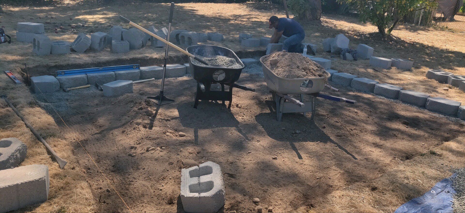 Person building a stone wall outdoors. Wheelbarrows, blocks, and tools are visible.