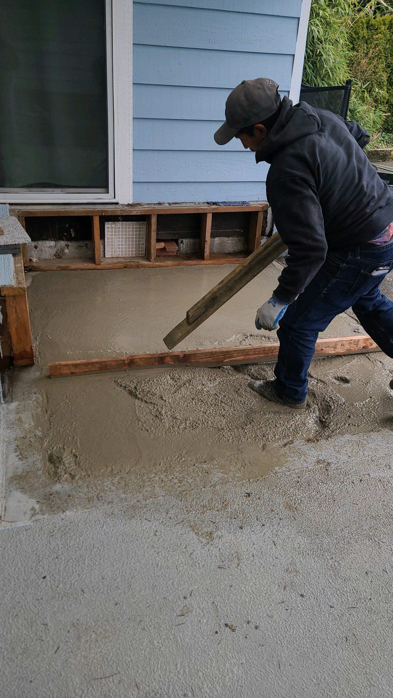 Man smoothing wet concrete with a screed tool on a porch; light blue building in background.
