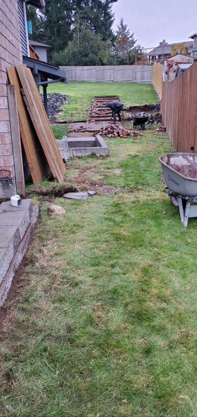 Backyard with grass, wooden fence, brick, a wheelbarrow and a grill.
